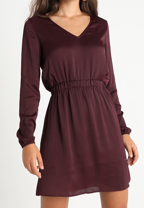 Mujer vestida con un vestido largo de manga larga, con escote en V, color burdeos oscuro, con cintura elástica y una longitud hasta la rodilla, contra un fondo liso.