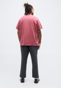 Άνδρας με κοτσίδες, φοράει ροζ πουκάμισο με κοντά μανίκια, σκούρο παντελόνι, λευκές κάλτσες και καφέ παπούτσια, στέκεται γυρισμένος ανάποδα σε απλό φόντο.