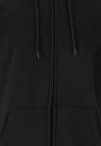 Mustassa vetoketjuhupparissa on pehmeä kangas, etutasku ja säädettävät nyörit. Saumat on ommeltu huomaamattomasti, eikä näkyviä logoja ole.