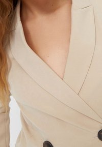 Béžový blazer s hlubokým V výstřihem, dvouřadým zapínáním a velkými černými knoflíky; hladká látka se jemným leskem.
