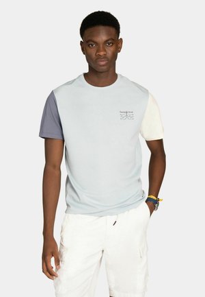 Giovane che indossa una t-shirt a blocchi di colore con maniche grigie e bianche e pantaloncini bianchi, in piedi con una mano in tasca.