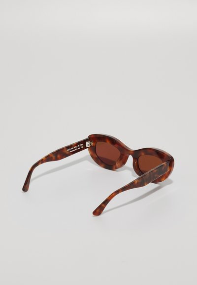 Lunettes de soleil en écaille de tortue avec des verres teintés marron, monture arrondie et branches fines. Comprend des détails de marque et de modèle sur la branche.