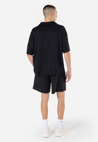 Chemise noire à manches courtes avec un col, portée sur un short en maille noir. La tenue est complétée par des baskets blanches et des chaussettes de sport.