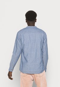 Ljusblå långärmad skjorta i chambray-material med krage, knappmanschetter och rundad fåll, bärs tillsammans med ljusrosa kordbyxor.