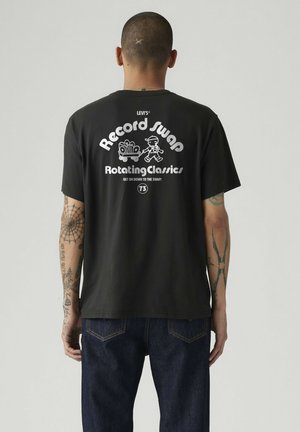 Uomo con braccia tatuate che indossa una t-shirt nera Levi's con testo bianco e cartone animato mentre tira un carrello pieno di dischi sul retro.