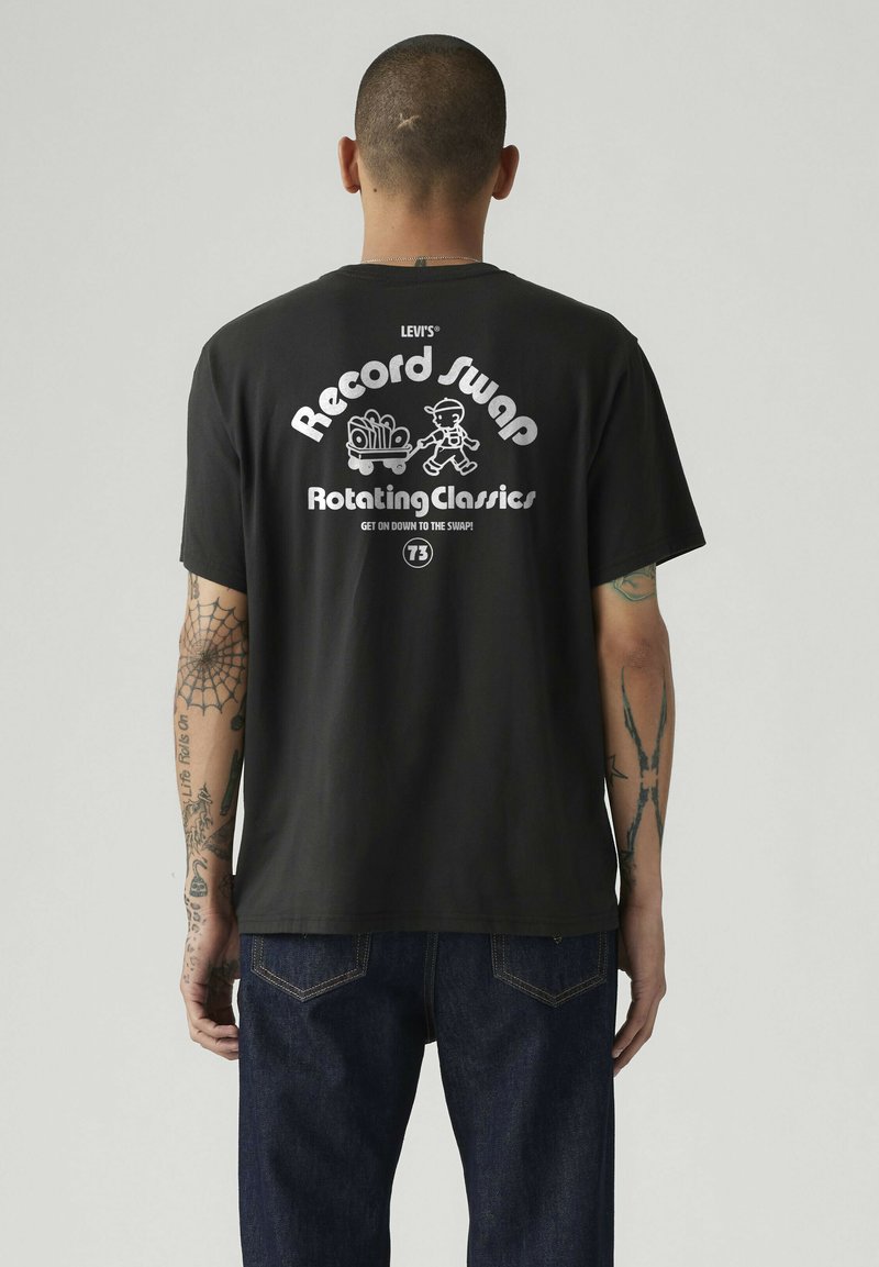 Hombre con brazos tatuados que lleva una camiseta negra de Levi's con texto blanco y un dibujo animado, tirando de un carro lleno de discos en la parte trasera.