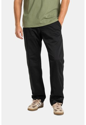 Man wearing black pants, green t-shirt, and beige sneakers with white stripes standing with one hand in pocket.