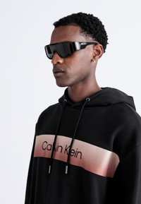Black hooded sweatshirt with a gradient pink band featuring "Calvin Klein" text. Model wears sleek black sunglasses with a sporty design.