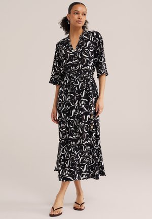 Kobieta w czarno-białej sukience midi z abstrakcyjnym wzorem, rękawami do łokcia i bocznym rozcięciem, zestawiona z czarnymi sandałami na obcasie.