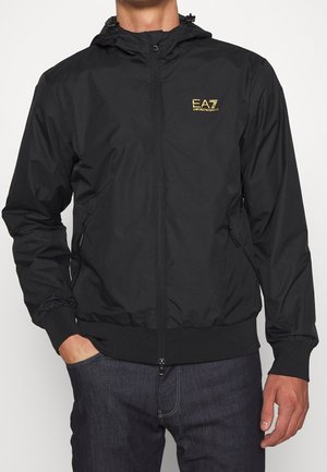 Mężczyzna w czarnej kurtce z kapturem na zamek błyskawiczny z żółtym logo "EA7 Emporio Armani" na piersi, w połączeniu z ciemnoniebieskimi dżinsami.