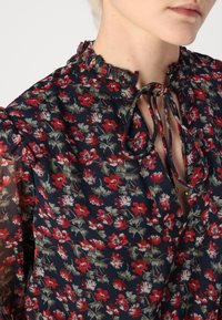 Blommig blus med volangkrage och knytning, med en marinblå bakgrund och röda samt gröna blommor, halvgenomskinliga ärmar.