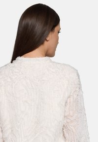 Weiße, strukturierte Jacke mit erhabenem, flauschigem Muster. Das Design wirkt weich und geschichtet, mit einem einfachen Ausschnitt und offenem Rücken.