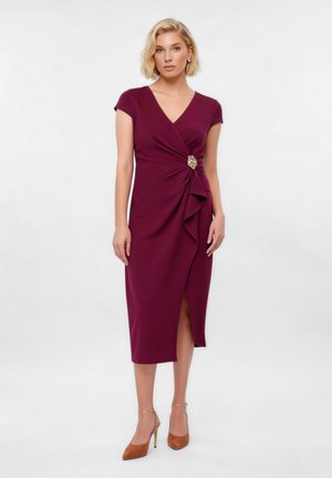 Kvinde iført knælængde, kortærmet bordeaux kjole med sideflæse og guldbroche, iført lysebrune højhælede sko, stående mod en lys baggrund.