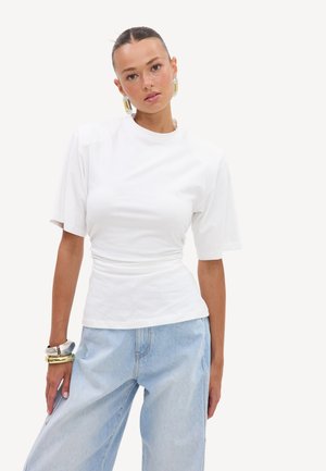 Femme aux cheveux tirés en arrière portant un top blanc moulant, un jean évasé bleu clair, de grandes boucles d’oreilles en créoles, et des bracelets en argent et en or.
