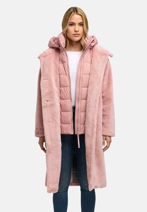 Frau trägt einen langen pinken Kunstpelzmantel über einer gesteppte pinke Jacke, weißem Hemd und blauen Jeans, steht vor weißem Hintergrund.