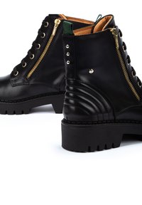Bottes de cheville en cuir noir avec des zips en or, bout rond, semelle chunky, détails à côtes sur le talon et accents métalliques sur les côtés.