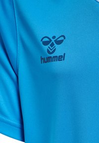 Chemise sportive bleue en tissu lisse et léger, arborant un logo "hummel" foncé bien visible et un motif abstrait au niveau de la poitrine.