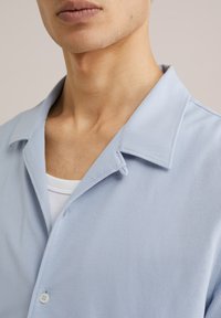 Camicia blu chiaro a maniche corte con tessuto testurizzato, colletto a punta e un singolo bottone davanti, indossata sopra una maglietta bianca.