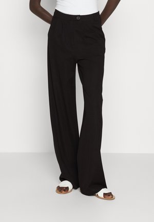 Persoon draagt zwarte, wijde broek met hoge taille, voorste knoop en zakken, gecombineerd met witte sandalen op een effen achtergrond.
