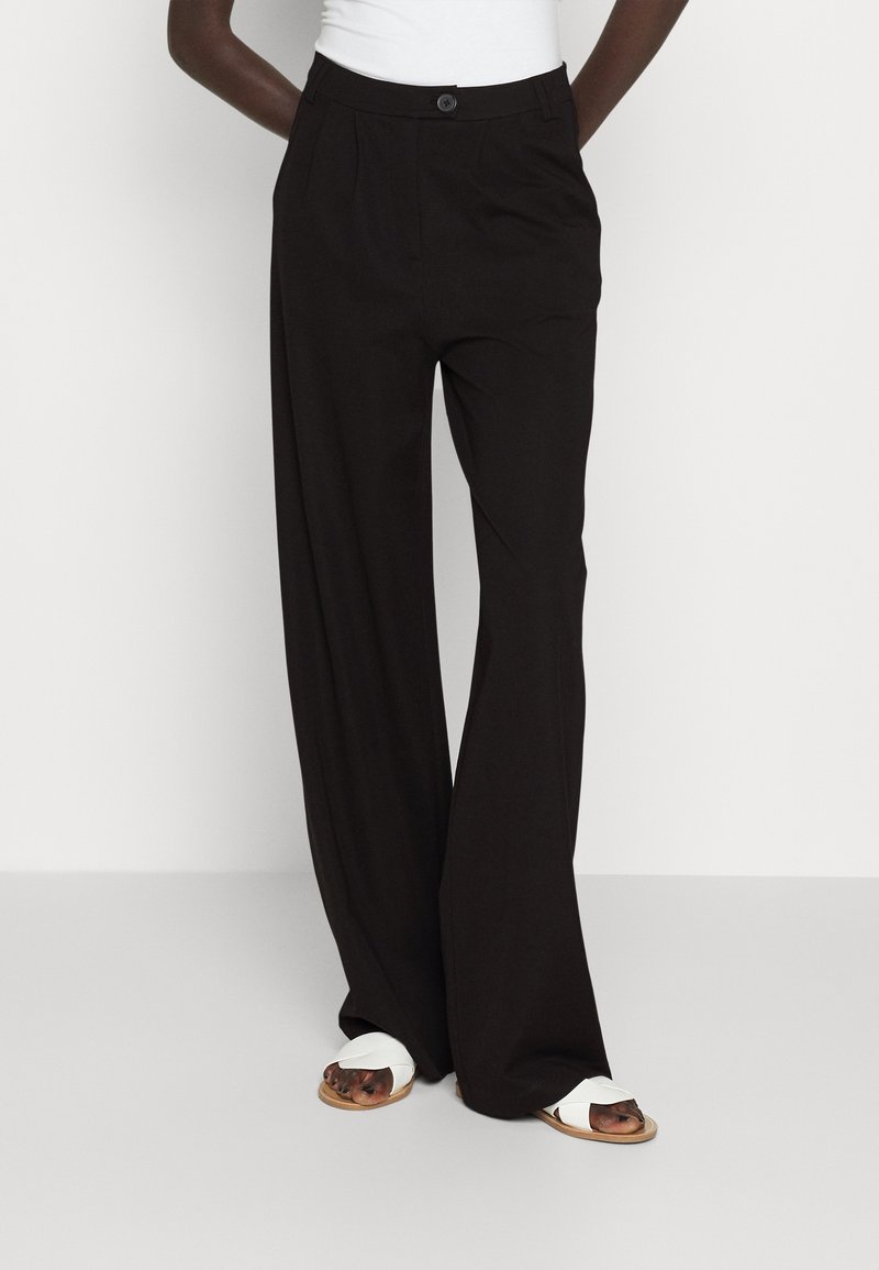 Personne portant un pantalon noir taille haute à jambes larges avec bouton sur le devant et poches, associé à des sandales blanches sur un fond uni.