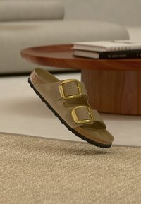 Μπεζ σανδάλι με δύο μεγάλες χρυσές αγκράφες και σόλα από φελλό που αιωρείται πάνω από μια υφασμάτινη μοκέτα σε ένα μοντέρνο καθιστικό.