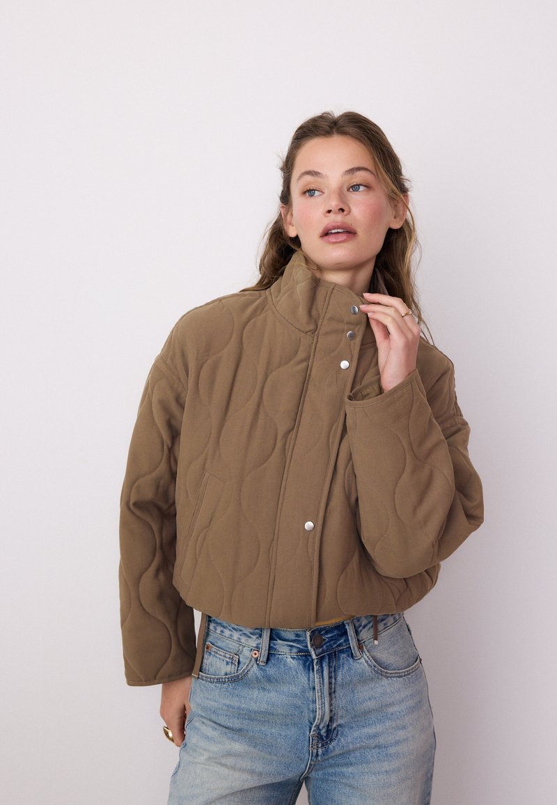 Žena si upravuje golier hnedej prešívanej bundy, nosí svetlomodré džínsy, na jednoduchom svetlom pozadí.