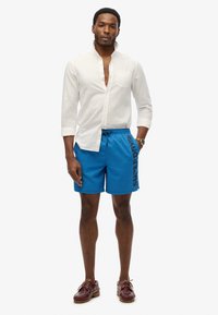 Shorts de bain bleus avec le logo "SUPERDRY" en noir, associés à une chemise en lin blanche à boutons et des chaussures en cuir marron.