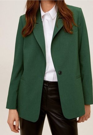 Femme portant un blazer vert par-dessus une chemise blanche à col et un pantalon en cuir noir, montrée du cou jusqu'à mi-cuisse sur un fond neutre.