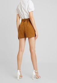 Bruna mockasins-shorts med hög midja och bältesdetalj, i kombination med vita klacksandaler. Tyget verkar mjukt och texturerat.