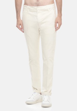 Uomo che indossa pantaloni aderenti di colore bianco sporco con orli a risvolto e sneakers bianche, in piedi su uno sfondo chiaro e uniforme.