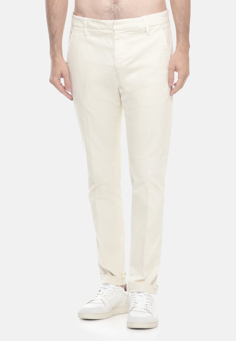 Uomo che indossa pantaloni aderenti di colore bianco sporco con orli a risvolto e sneakers bianche, in piedi su uno sfondo chiaro e uniforme.