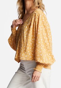 Blouse jaune moutarde avec imprimé floral blanc, manches bouffantes et poignets froncés. Tissu léger et fluide avec un décolleté en V profond.