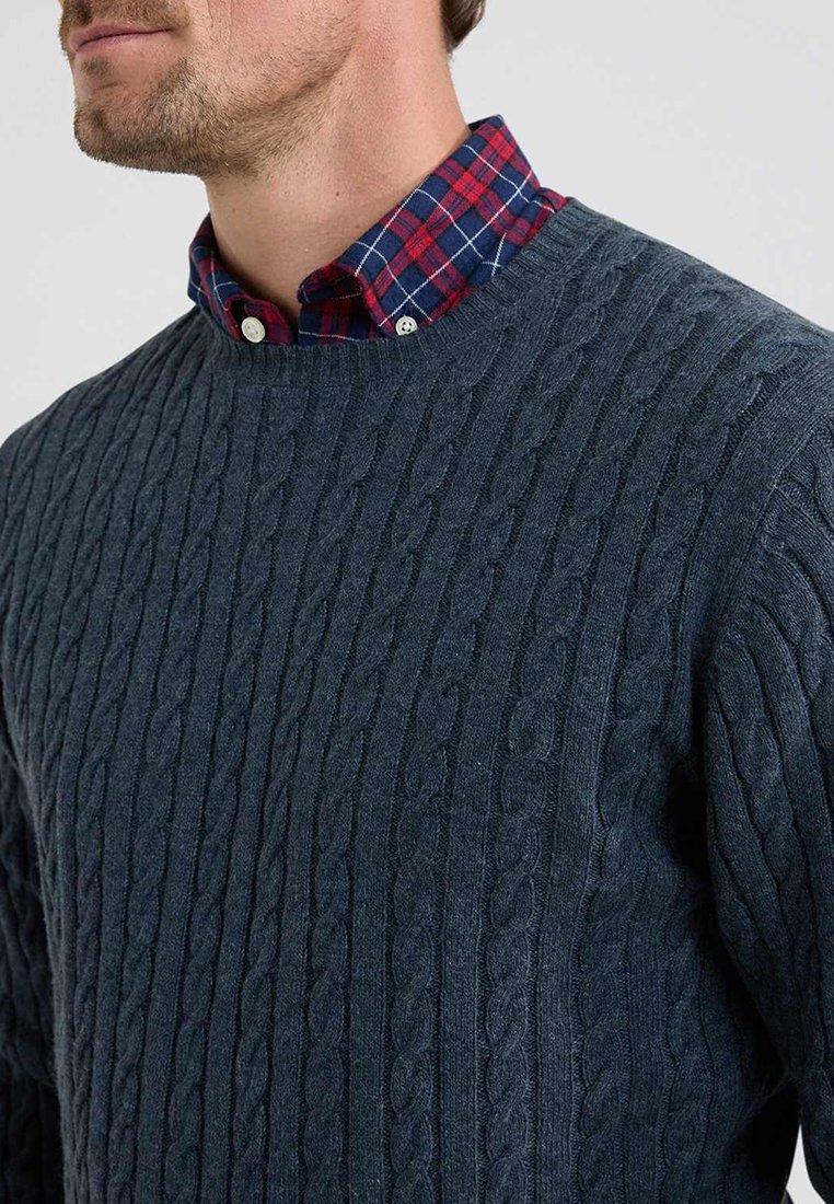 Dik marineblauw kabelgebreide trui met een ronde hals, gedragen over een rood en marineblauw geruit shirt. De stof lijkt zacht met gestructureerde patronen.