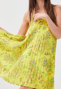 Robe fleurie jaune avec des bretelles fines, en tissu léger, texture plissée, ornée de motifs complexes dans des teintes de vert, orange et bleu.