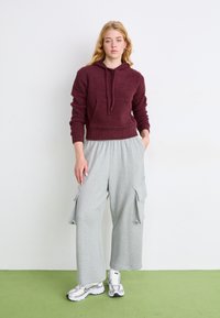 Bordo puhasti hoodie s sprednjo žepom, v kombinaciji z svetlo sivo širokima hlačama s stranskimi žepi. Obutev: srebrni športni copati.