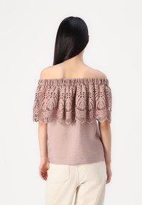 Blusa off-the-shoulder in rosa pallido con sovrapposizione di pizzo che presenta intricati motivi a taglio. Tessuto morbido, leggermente strutturato, con maniche a sbuffo.