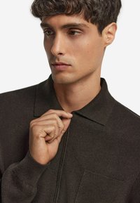 Mörkbrun stickad polo t-shirt med krage och ficka, med ribbade ärmslut och en strukturerad tyg. Modellen justerar kragen.
