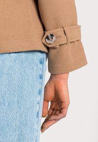 Tæt siddende tan uldjakke med bred manchet, der har en rund, klar knap, parret med lys blå denim jeans. Blødt, tekstureret materiale.