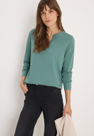Top teal a maniche lunghe con scollatura a V, abbinato a pantaloni neri. Il tessuto presenta una leggera texture. Il modello tiene un indumento color crema.