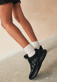 Black athletic shoes with a textured finish, accented by a white grid pattern, paired with white ribbed socks.