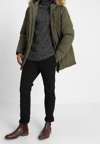 Olivgrüne Daunenjacke mit fellgefütterter Kapuze, grauer Strickpullover, schwarze Hose und braune Lederchelsea-Boots. Lässiges Winteroutfit.