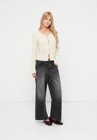 Cardigan en maille crème à boutons, avec un motif texturé, associé à un jean noir délavé, taille haute et jambes larges.