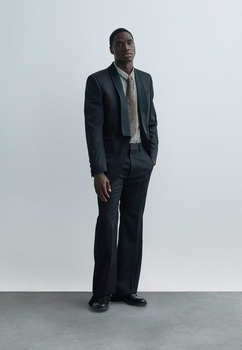 Costume noir avec un revers en satin vert, assorti à une chemise à motif et une cravate marron. Des chaussures noires complètent le look. Fond gris simple.