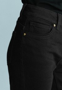 Gros plan d'une poche de jean noire avec des rivets dorés et des détails de couture, portée avec un haut sombre.