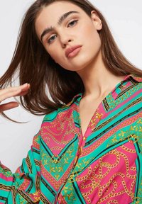 Camisa de botones colorida con un patrón geométrico en rosa, azul verdoso y negro, con acentos dorados y diseños de cadenas en una tela suave.