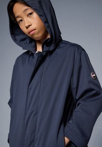 Navy blue hooded jacket made from smooth fabric; features a button closure, slightly oversized fit, and a logo patch on the sleeve.
