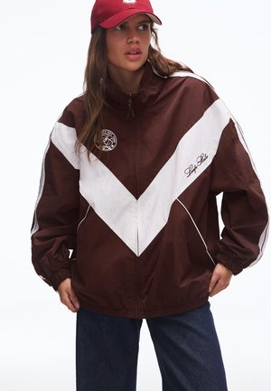 Žena oblečená v hnedom a bielom sakom so vzorom chevron, s červenou šiltovkou a tmavými džínsami, pozrieva sa mierne do strán s rukami v vreckách.