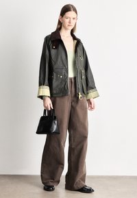 Roheline vahatatud jakk pruunide krae, kuldsete detailide ja triibuliste varrukaservadega, koos pruunide laiade pükstega ja väikese musta käekotiga.