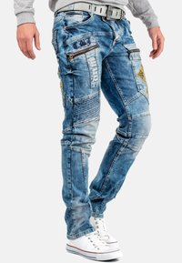 Jeans in denim blu sbiadito con diverse tasche con zip, pannelli testurizzati e cuciture decorative. Indossati con sneaker bianche.