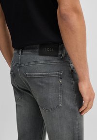 Jeans de mezclilla gris oscuro con una textura suave, que presentan un parche de cuero en la cintura y un acento plateado en el bolsillo trasero.